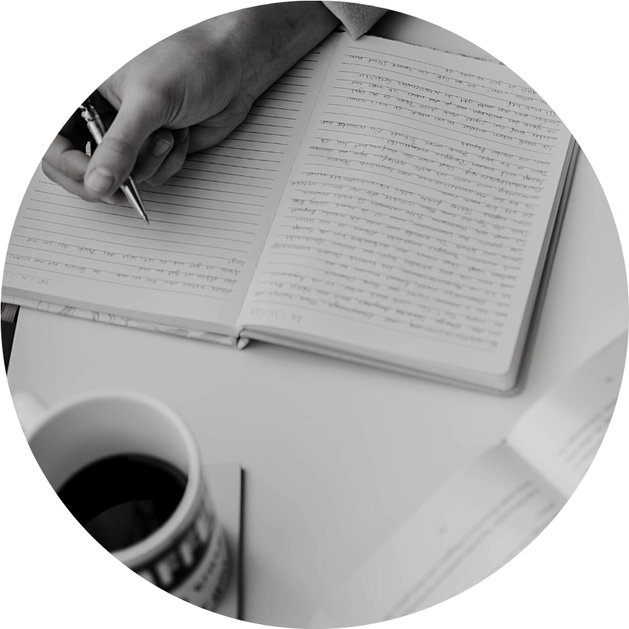 The image is a circle. A hand holds a pen, hovering over the journal she's writing in. Across from her is a mug with the letters FFE showing and dark coloured liquid inside.