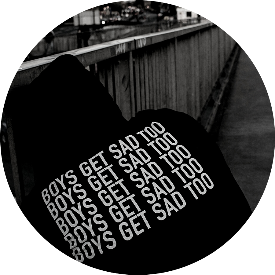 Close up of the back of a black hoodie worn by somone whose face cannot be seen. On the back of the hoodie is the phrase "Boys get sad too" repeated 5 times.