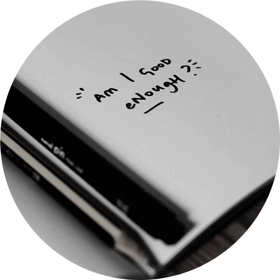 Close up shot of a notebook with the words "am I good enough?" written on it, there is a pen and a pencil in the crook of the spine