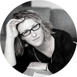 A white woman is lying on a grey couch, facing the camera. She is wearing a black shirt and black glasses.