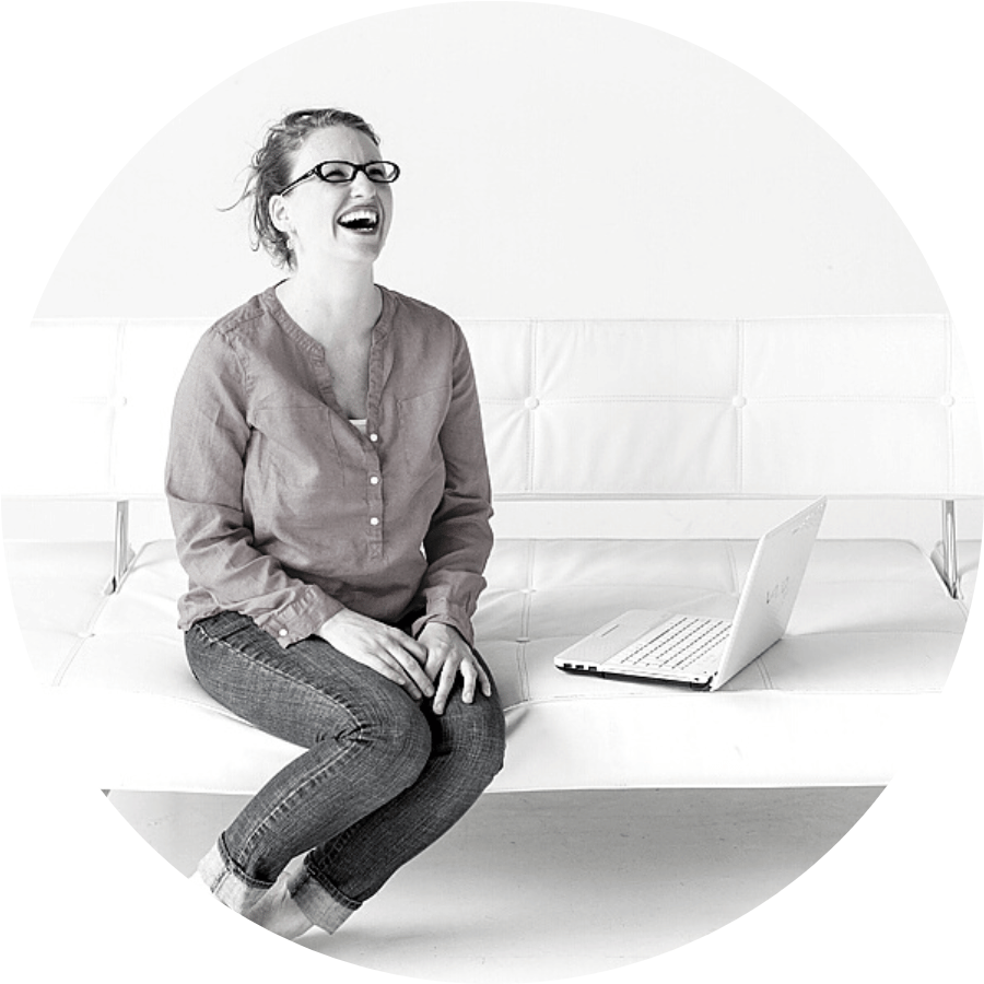 Parrish Wilson, a white woman, is sitting on a white couch laughing. In front of her is an open laptop. The image is in a monotone gray hue.