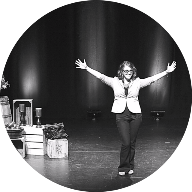 Parrish Wilson, a white woman, is standing on a stage with her arms raised and spread in joy with a big smile on her face.